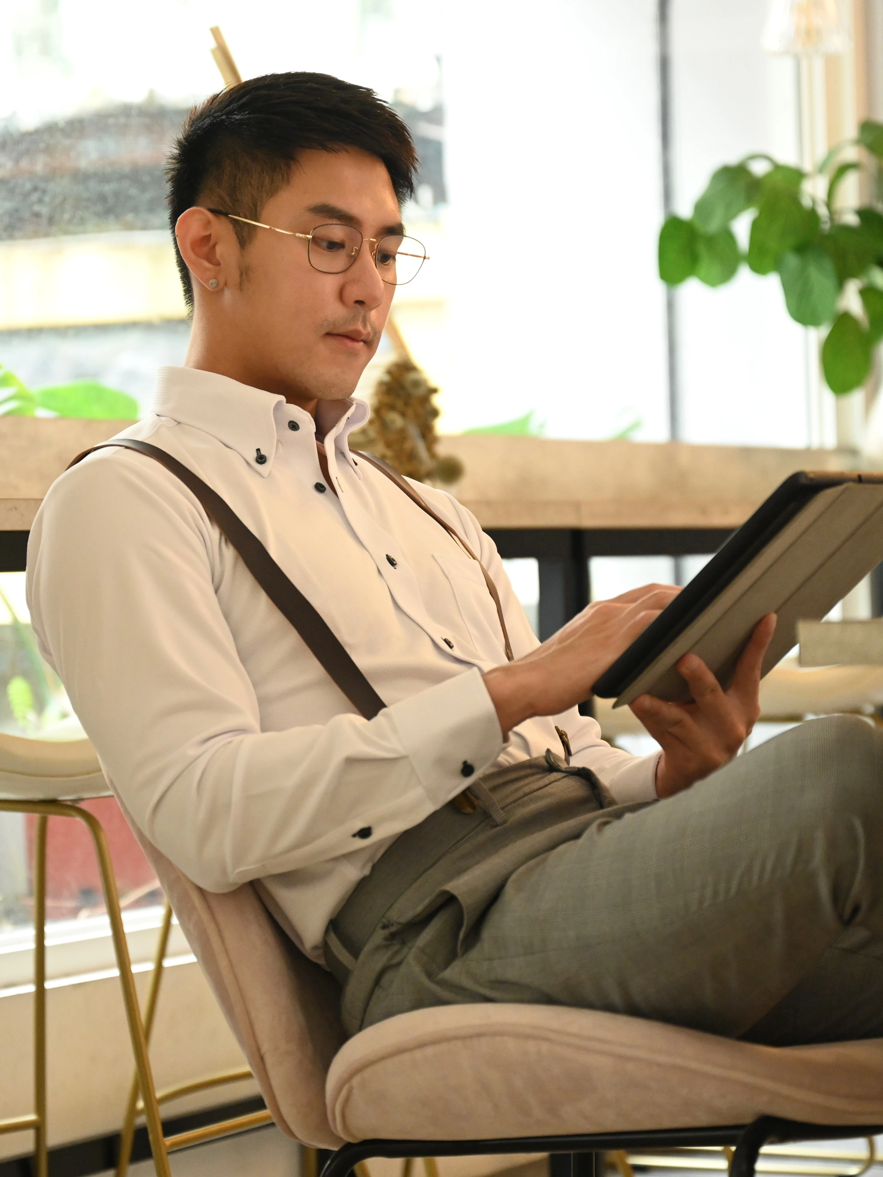 戴眼鏡男子穿著白色長袖襯衫與深色吊帶,坐在咖啡廳椅子上專注操作平板電腦,展現斯文商務休閒穿搭。